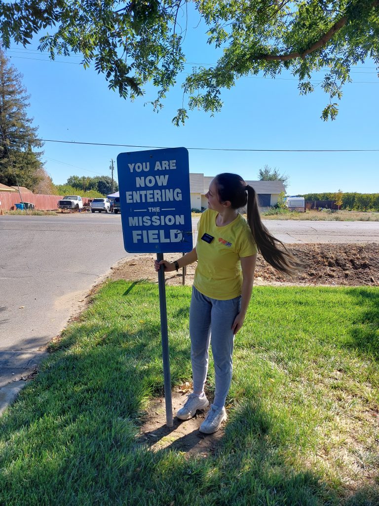 LDS Missionary standing with "You are now entering the mission field" sign.